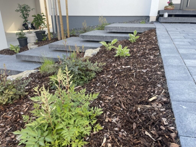 Schwebende, graue Treppe zu einem Eingangsbereich, zu dem ebenso ein gepflasteter Weg führt