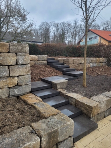 Eine anthrazitfarbene, schwebende Treppe mit 12 versetzten Stufen. Eine Natursteinmauer, die zwei Ebenden schafft.